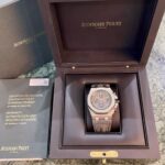 Audemars Piguet watch in its black display box with gold hardware, opened to reveal the timepiece and warranty booklet nearby.