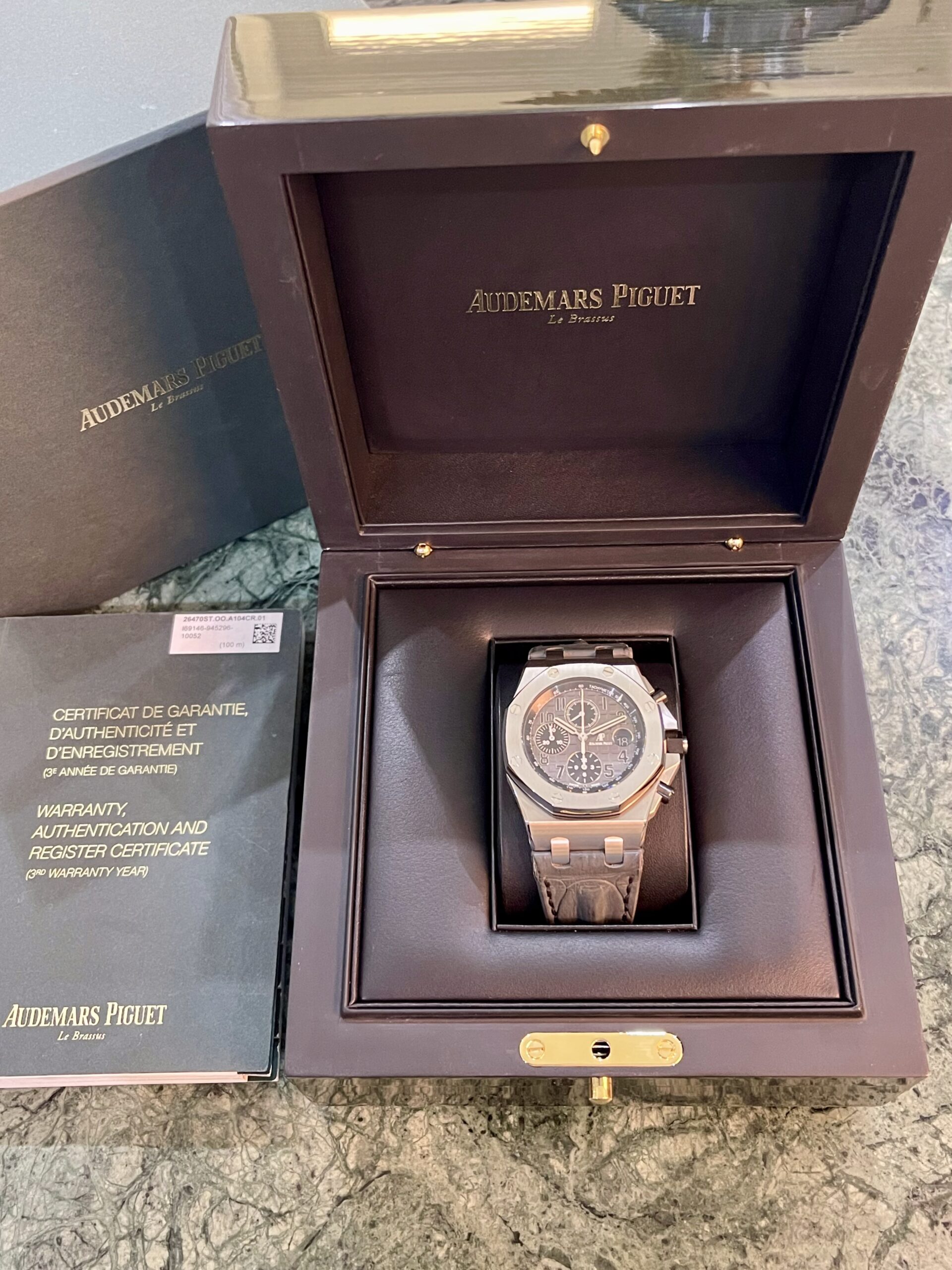 Audemars Piguet watch in its black display box with gold hardware, opened to reveal the timepiece and warranty booklet nearby.