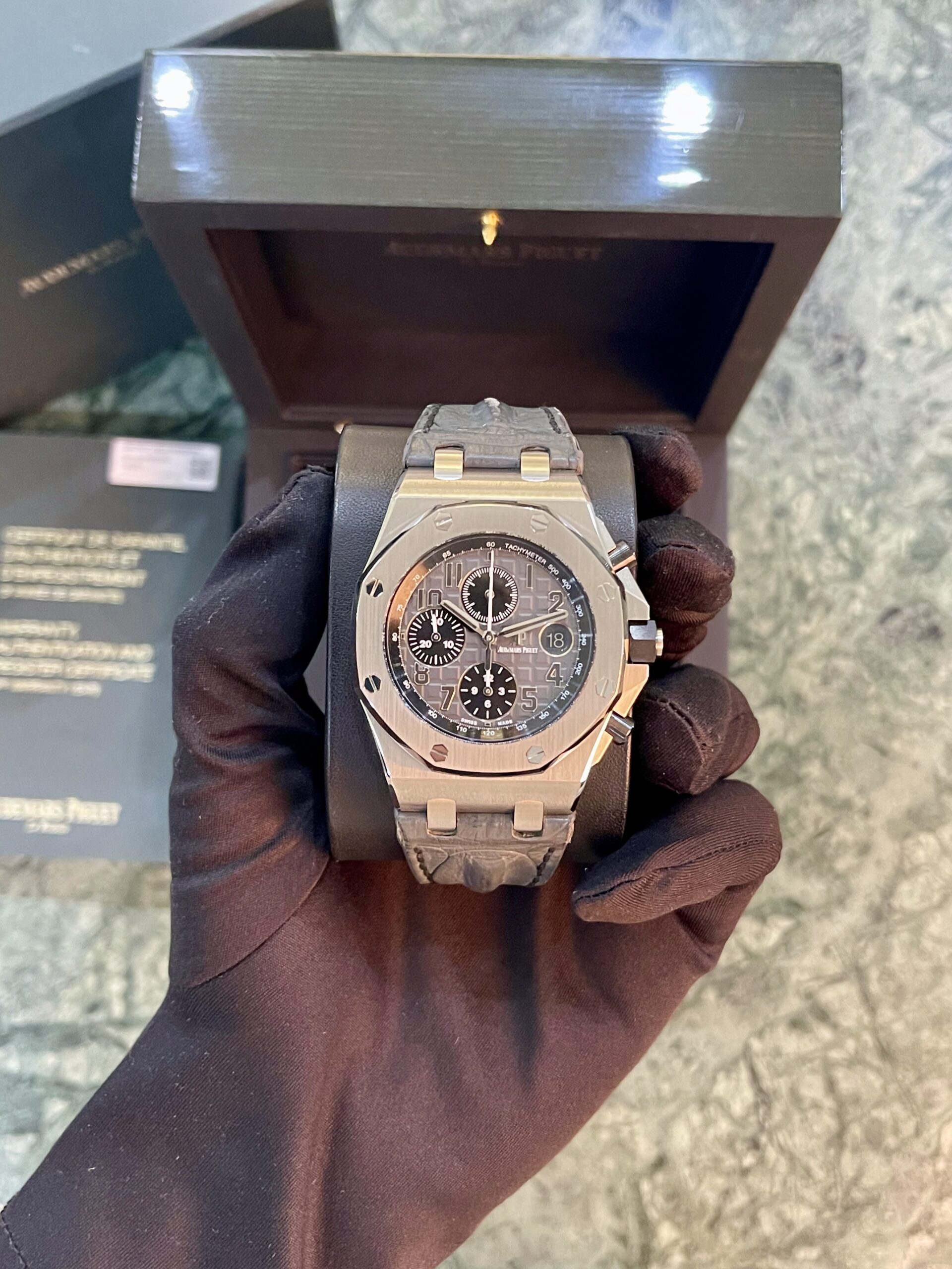 Stainless steel chronograph wristwatch held by a gloved hand, with a dark presentation box in the background.