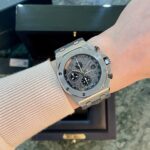 Wrist wearing a silver stainless steel chronograph watch with black dial, subdials, and date window, on a marble surface with an open box in the background.