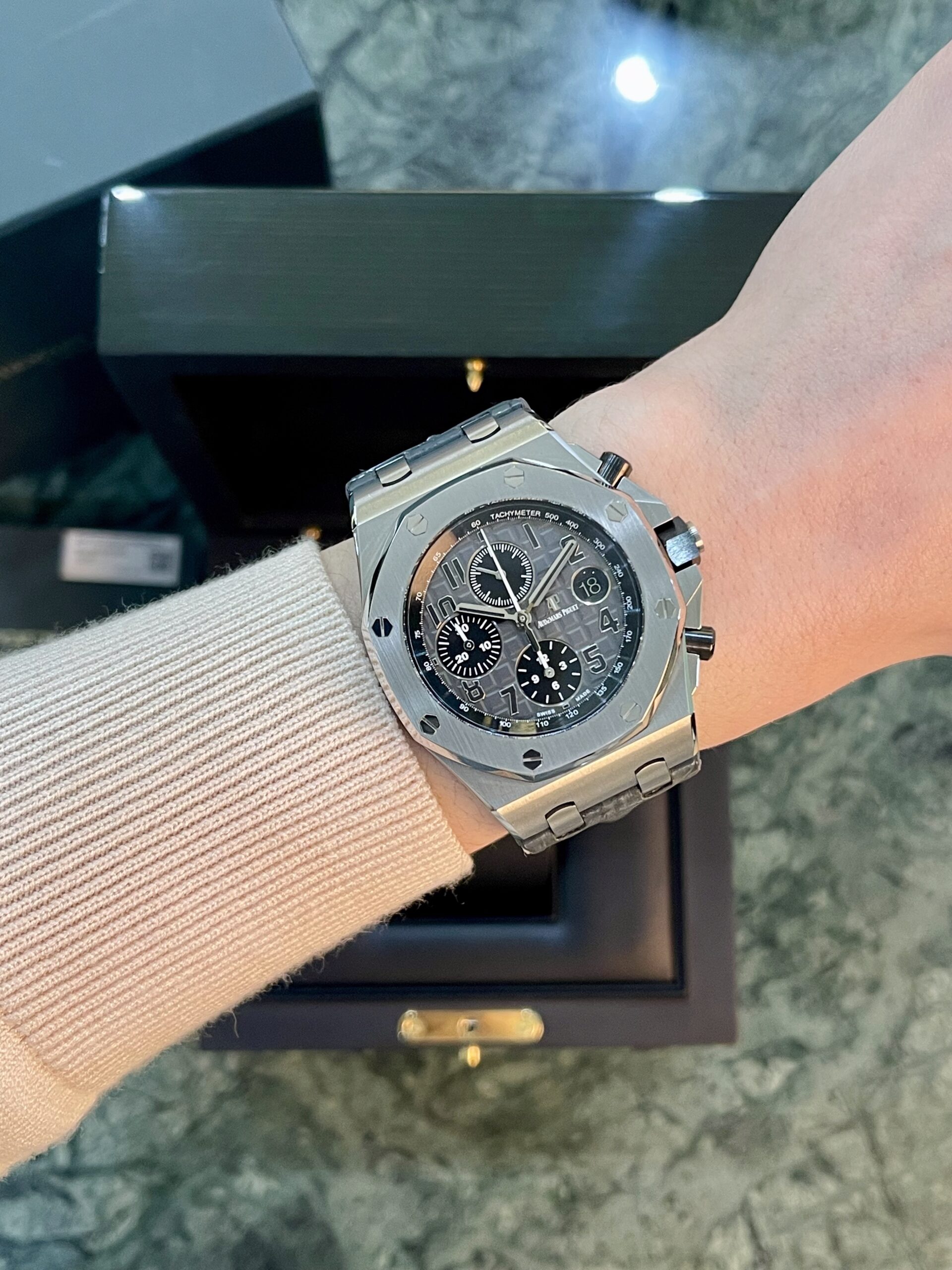 Wrist wearing a silver stainless steel chronograph watch with black dial, subdials, and date window, on a marble surface with an open box in the background.