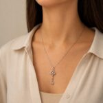 Silver key pendant necklace resting on a beige blouse, close-up of the jewelry design with a delicate chain and decorative bow-like top.