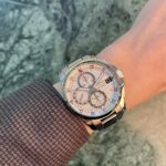 Stainless steel chronograph wristwatch on a wrist with a textured brown sleeve, white dial and multiple subdials.