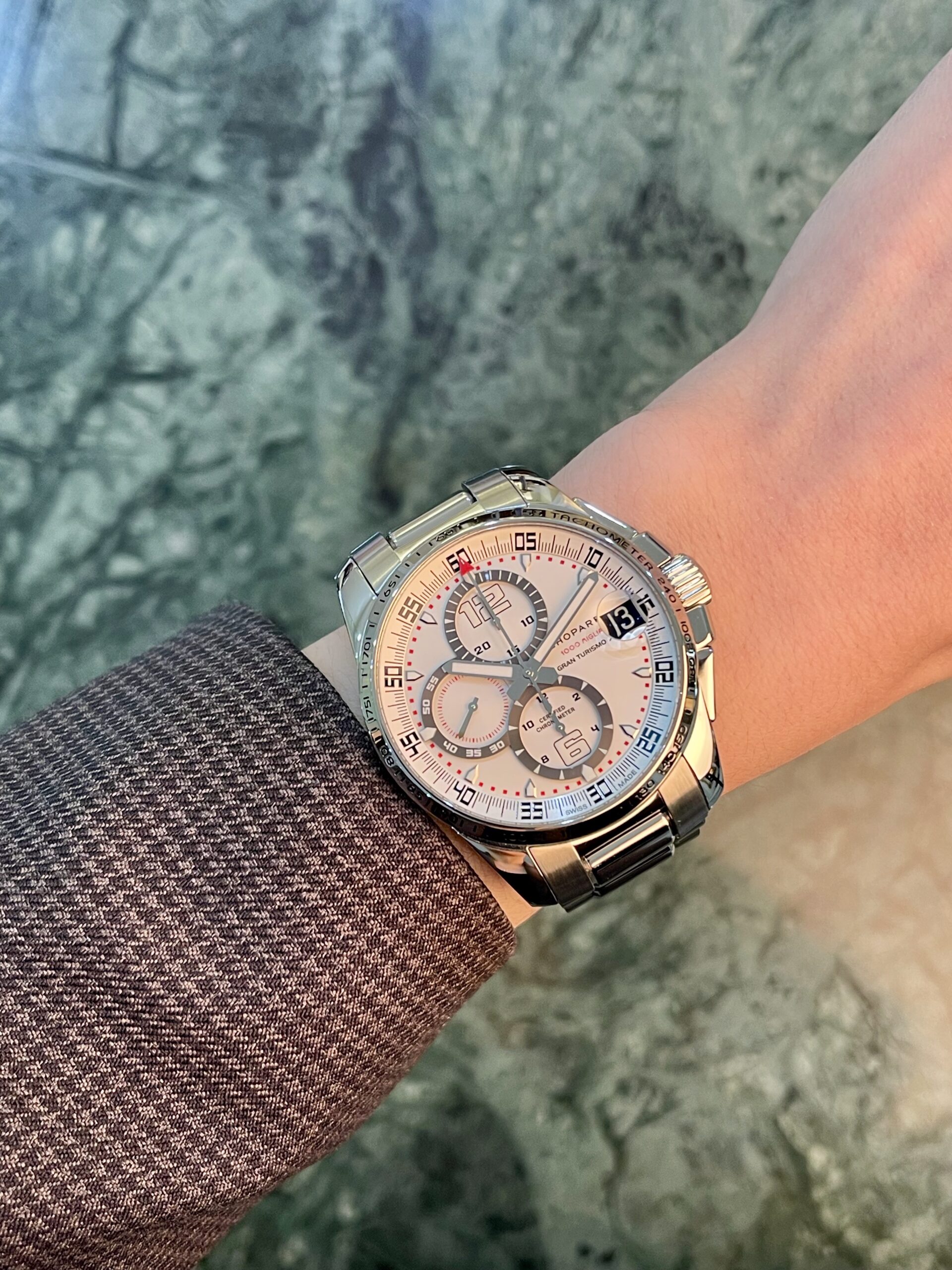 Stainless steel chronograph wristwatch on a wrist with a textured brown sleeve, white dial and multiple subdials.