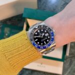 Wrist wearing a silver Rolex GMT-Master II with a blue and black bezel, on a yellow knit sleeve with a Rolex box in the background.