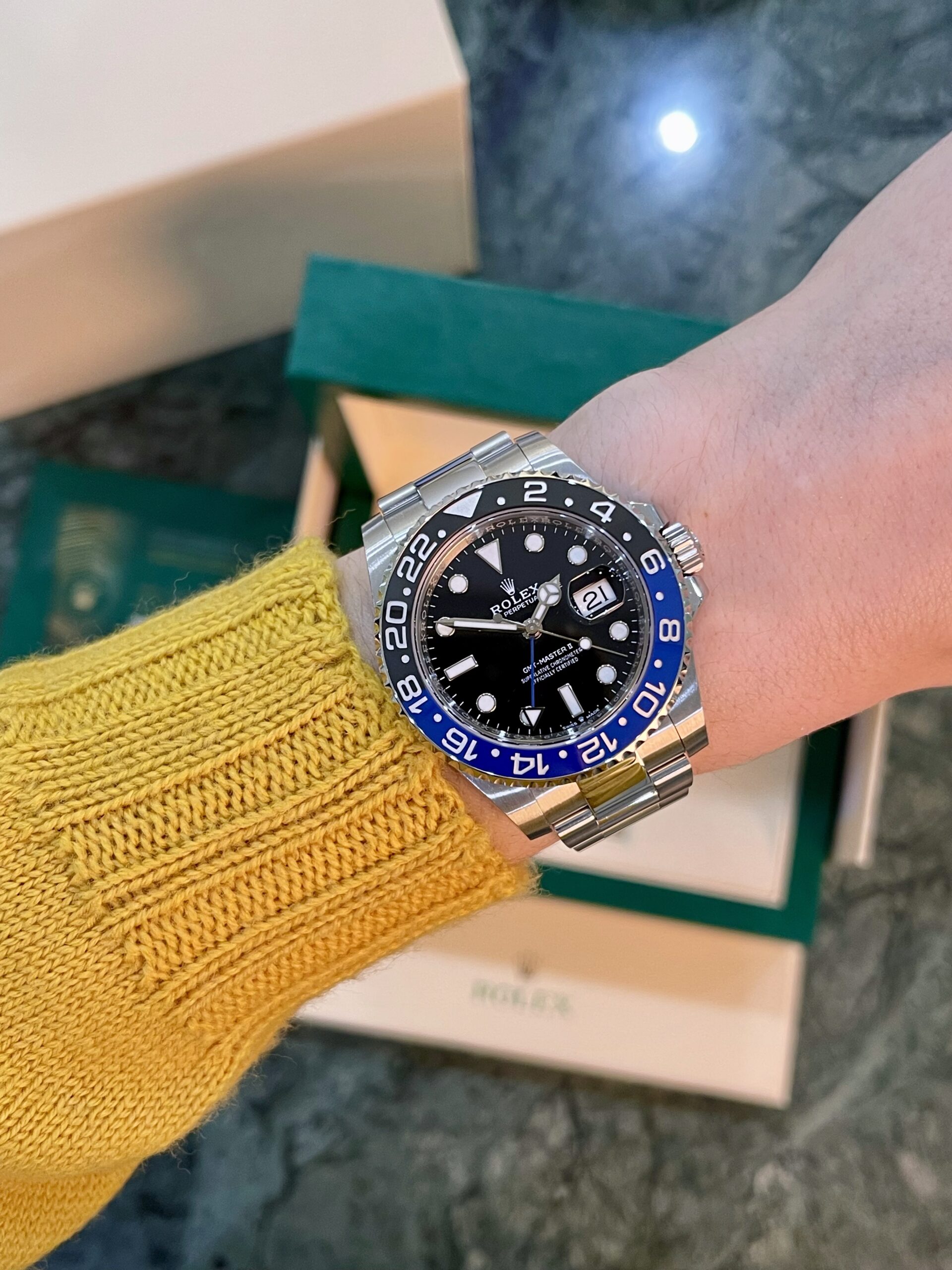 Wrist wearing a silver Rolex GMT-Master II with a blue and black bezel, on a yellow knit sleeve with a Rolex box in the background.