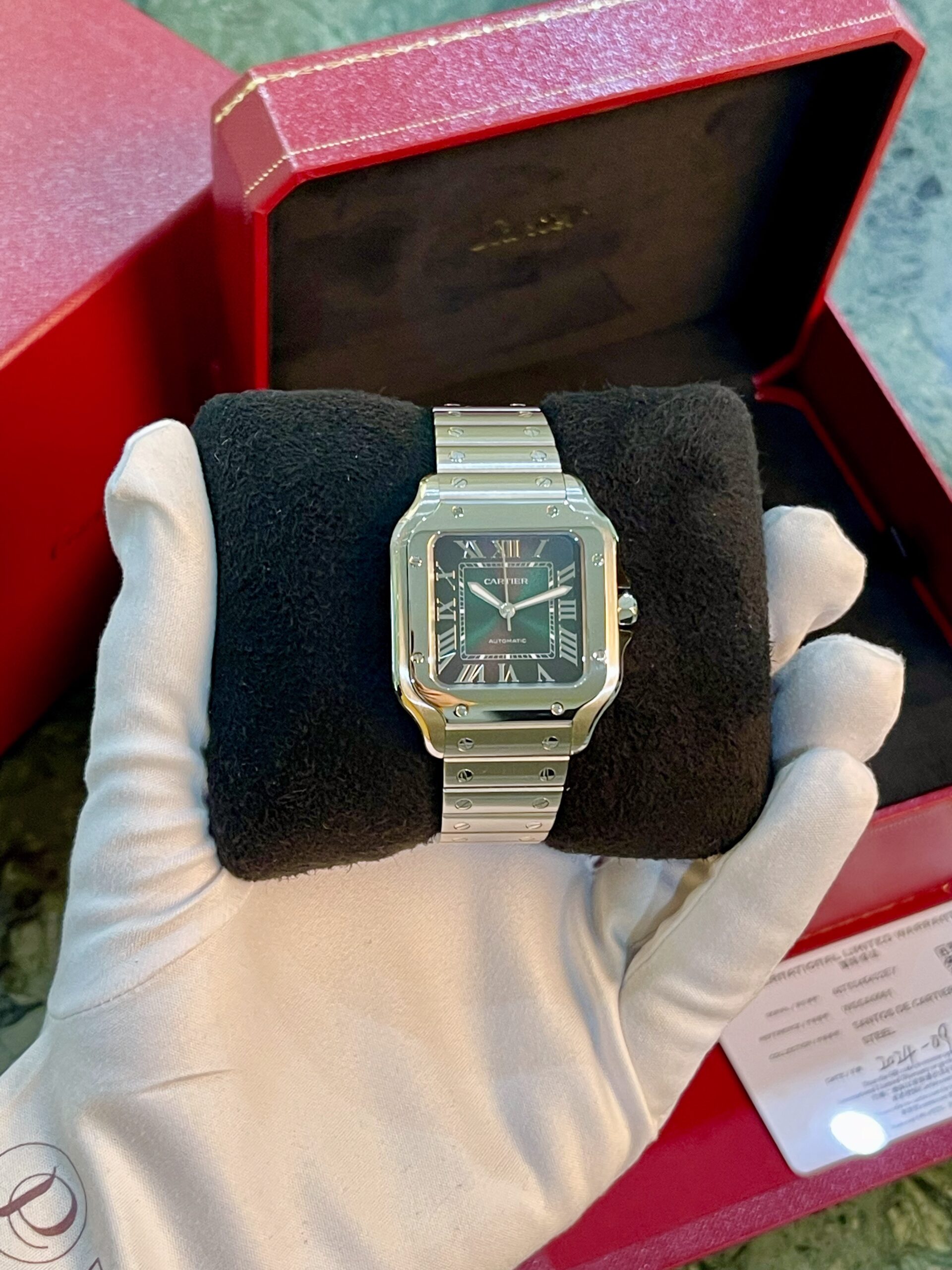 Cartier Santos stainless-steel watch with Roman numerals on a square face, displayed on a black cushion inside a red presentation box.