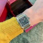 Close-up of a silver Cartier wristwatch on a yellow knit sleeve, with a red open box in the background.
