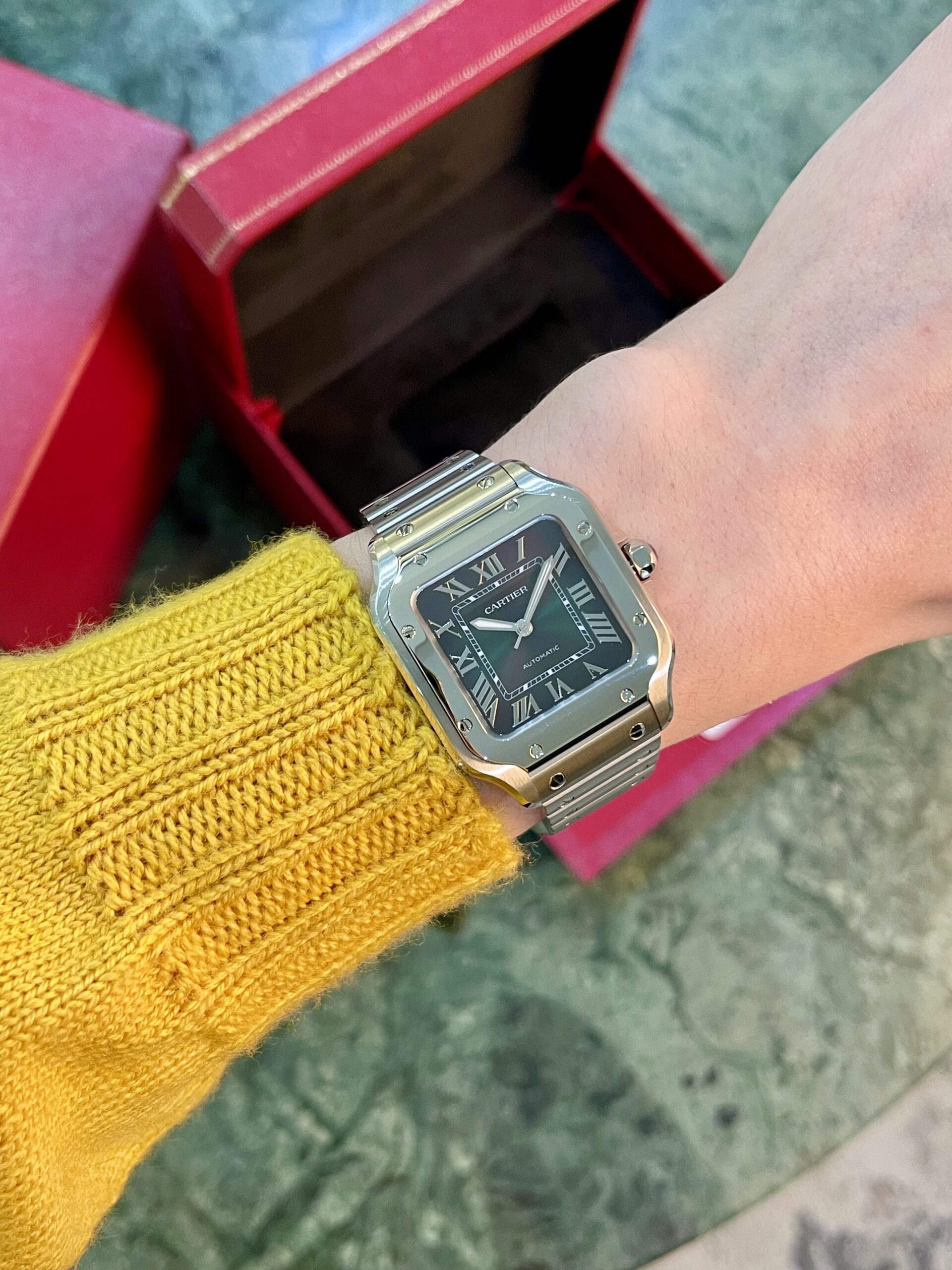 Close-up of a silver Cartier wristwatch on a yellow knit sleeve, with a red open box in the background.