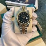 Rolex Datejust wristwatch with a two-tone gold and steel bracelet and diamond hour markers held by a gloved hand.