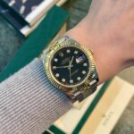 Two-tone Rolex Datejust on a wrist with a black dial, diamond hour markers, gold fluted bezel, and a two-tone bracelet; date window visible at 3 o'clock.
