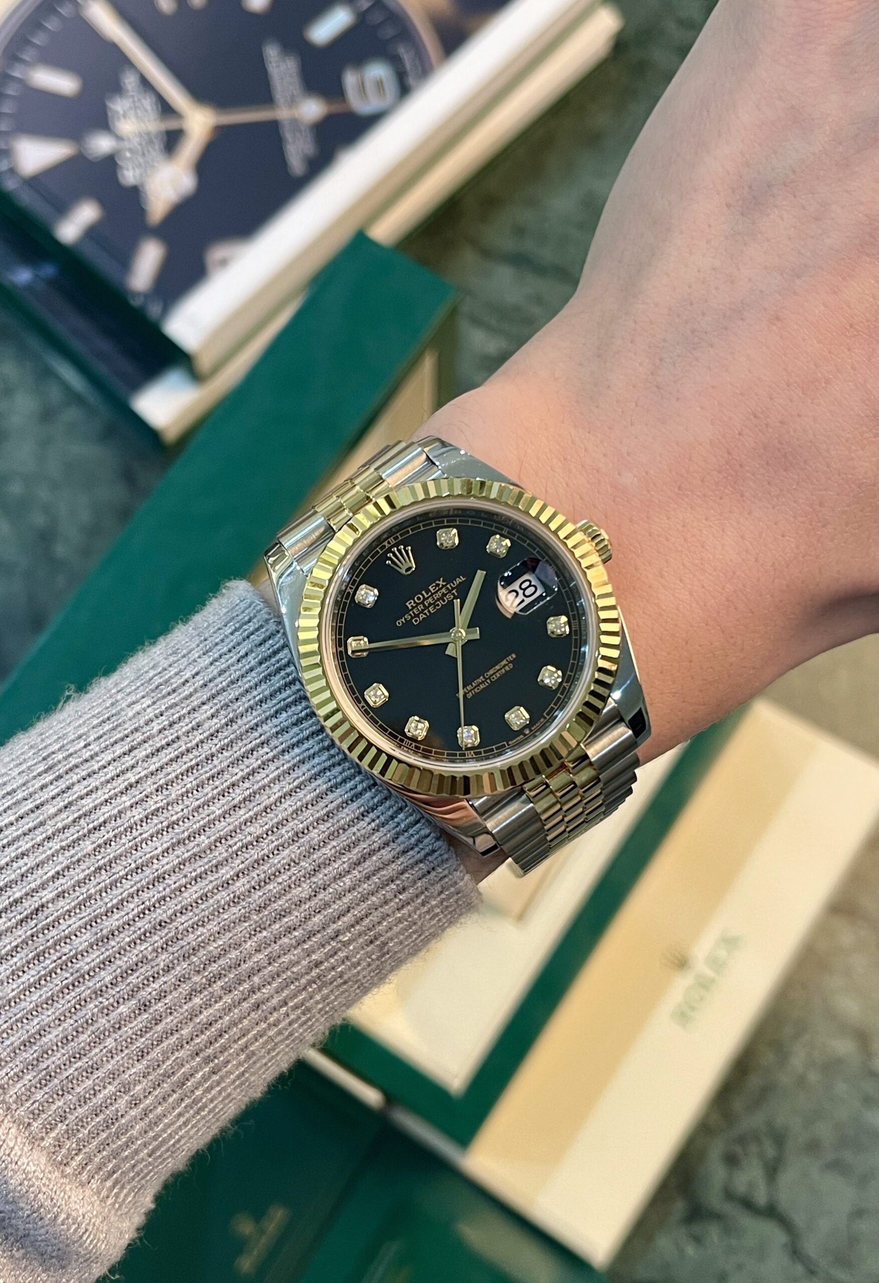 Two-tone Rolex Datejust on a wrist with a black dial, diamond hour markers, gold fluted bezel, and a two-tone bracelet; date window visible at 3 o'clock.