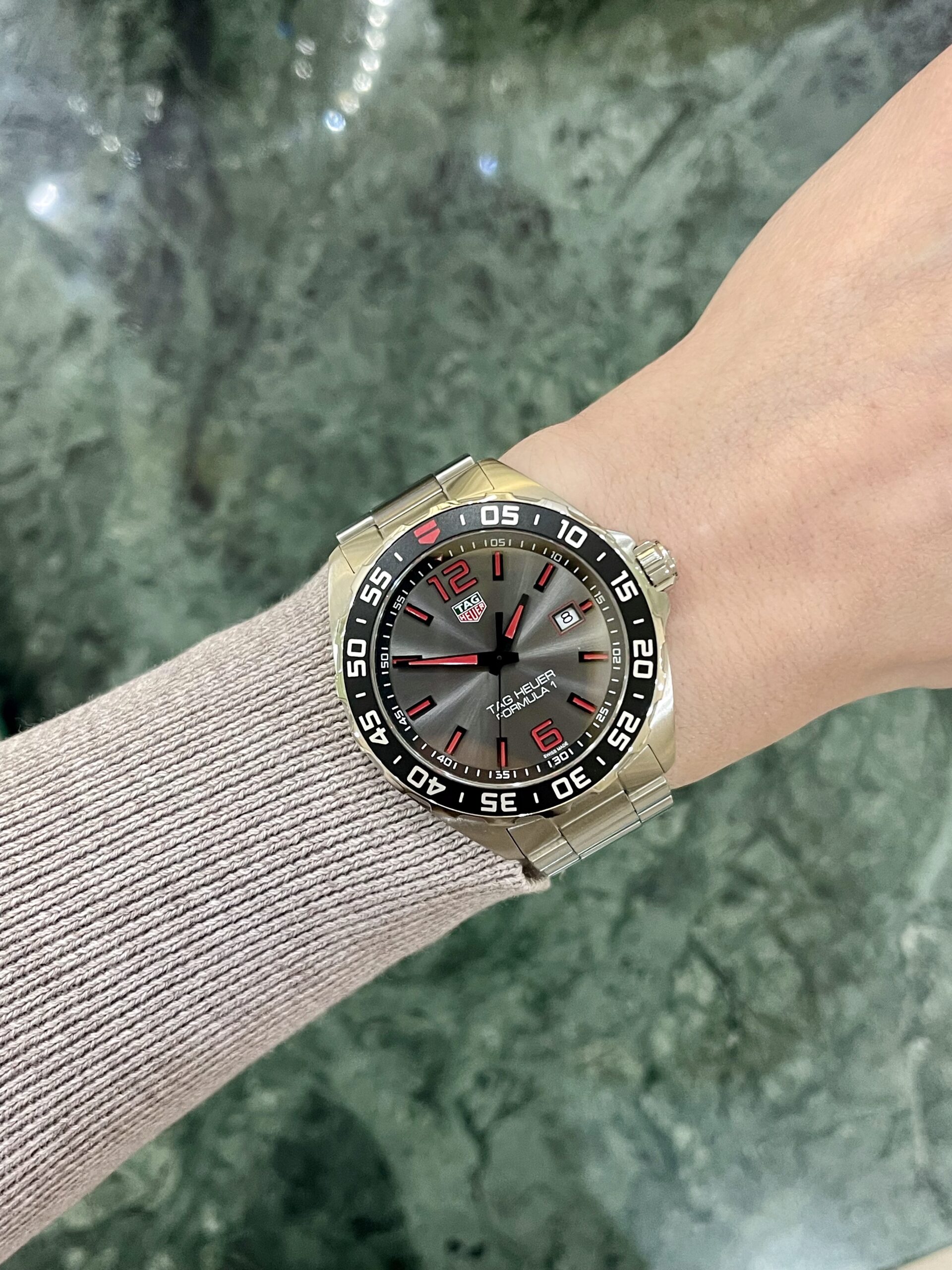 Stainless steel TAG Heuer Formula 1 watch on a wrist with a beige knit sleeve, against a green marble-like background.
