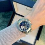Wrist wearing a stainless steel Zenith chronograph watch with a blue dial, against a beige sweater and open watch box in the background