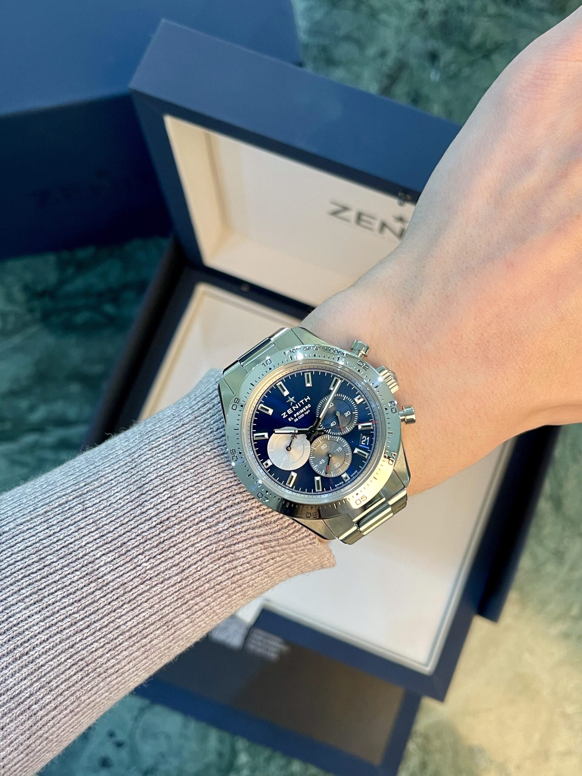 Wrist wearing a stainless steel Zenith chronograph watch with a blue dial, against a beige sweater and open watch box in the background