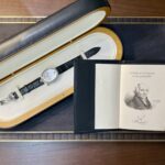 Wristwatch with black leather strap in an open presentation case beside a certificate booklet labeled Certificate d’origine et de garantie and a light interior lining.