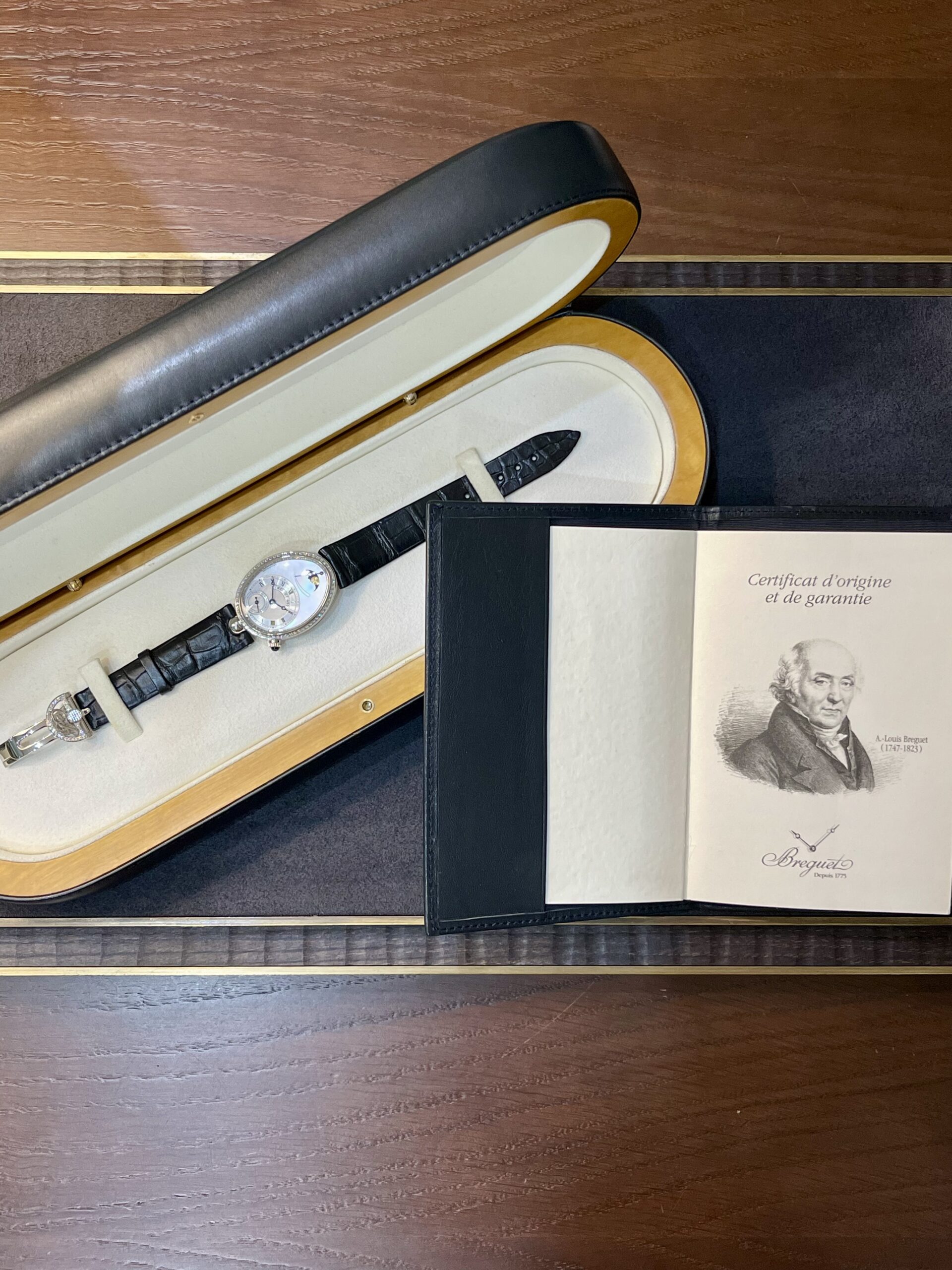 Wristwatch with black leather strap in an open presentation case beside a certificate booklet labeled Certificate d’origine et de garantie and a light interior lining.