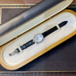Wristwatch with a black leather strap displayed inside a cream-lined wooden box case.