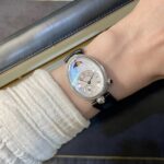 Close-up of a wrist wearing a silver-toned watch with a diamond-studded bezel and moon phase dial, paired with a black strap and a light knit sweater sleeve.