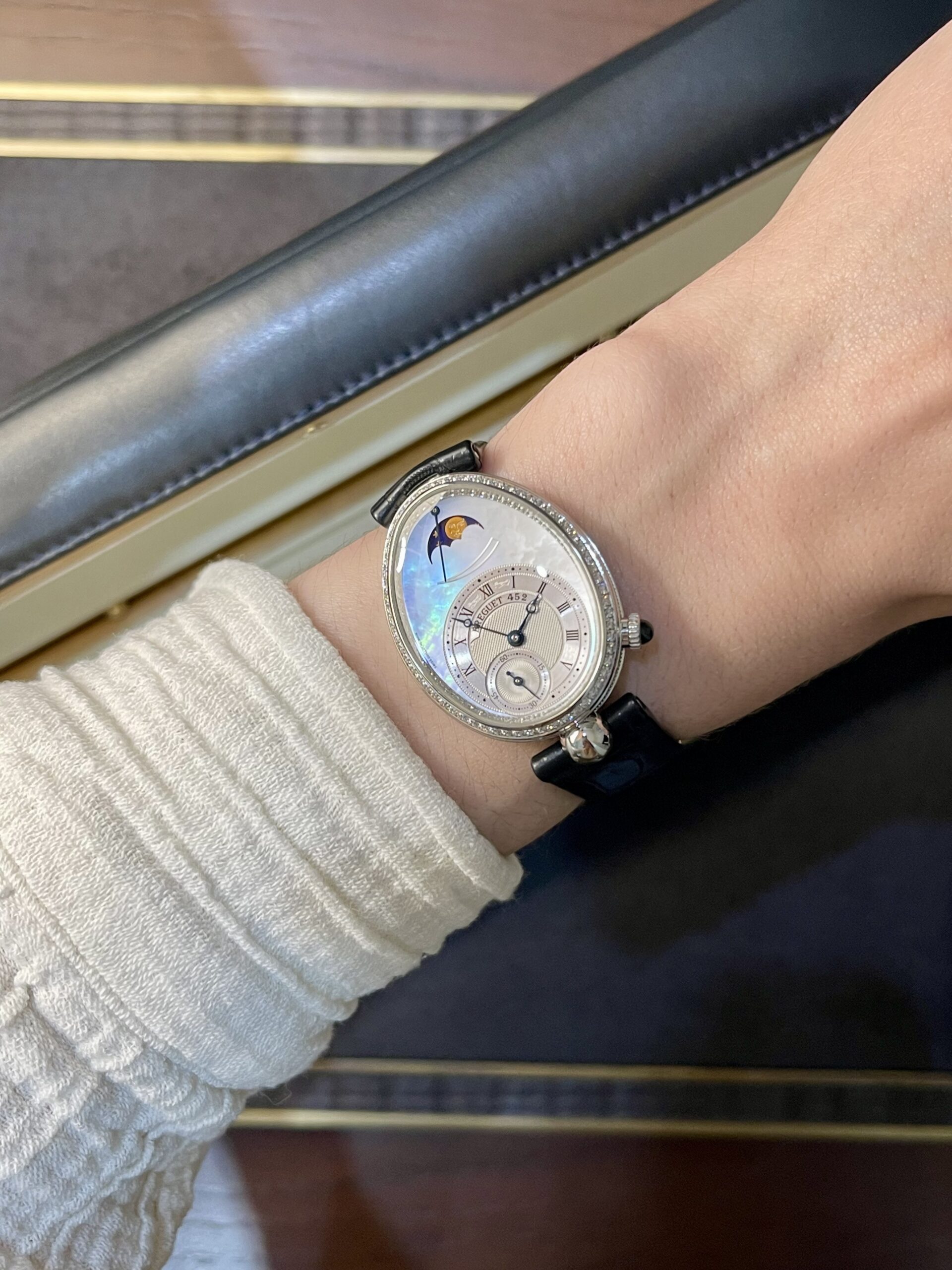 Close-up of a wrist wearing a silver-toned watch with a diamond-studded bezel and moon phase dial, paired with a black strap and a light knit sweater sleeve.