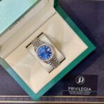 Luxury wristwatch with a blue dial and diamond-studded bezel in a beige display box inside a green presentation box outside.