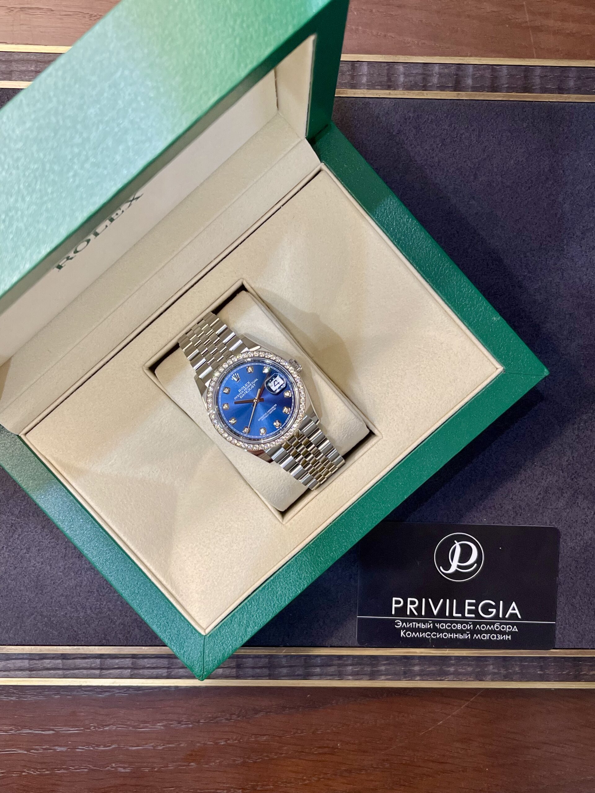 Luxury wristwatch with a blue dial and diamond-studded bezel in a beige display box inside a green presentation box outside.