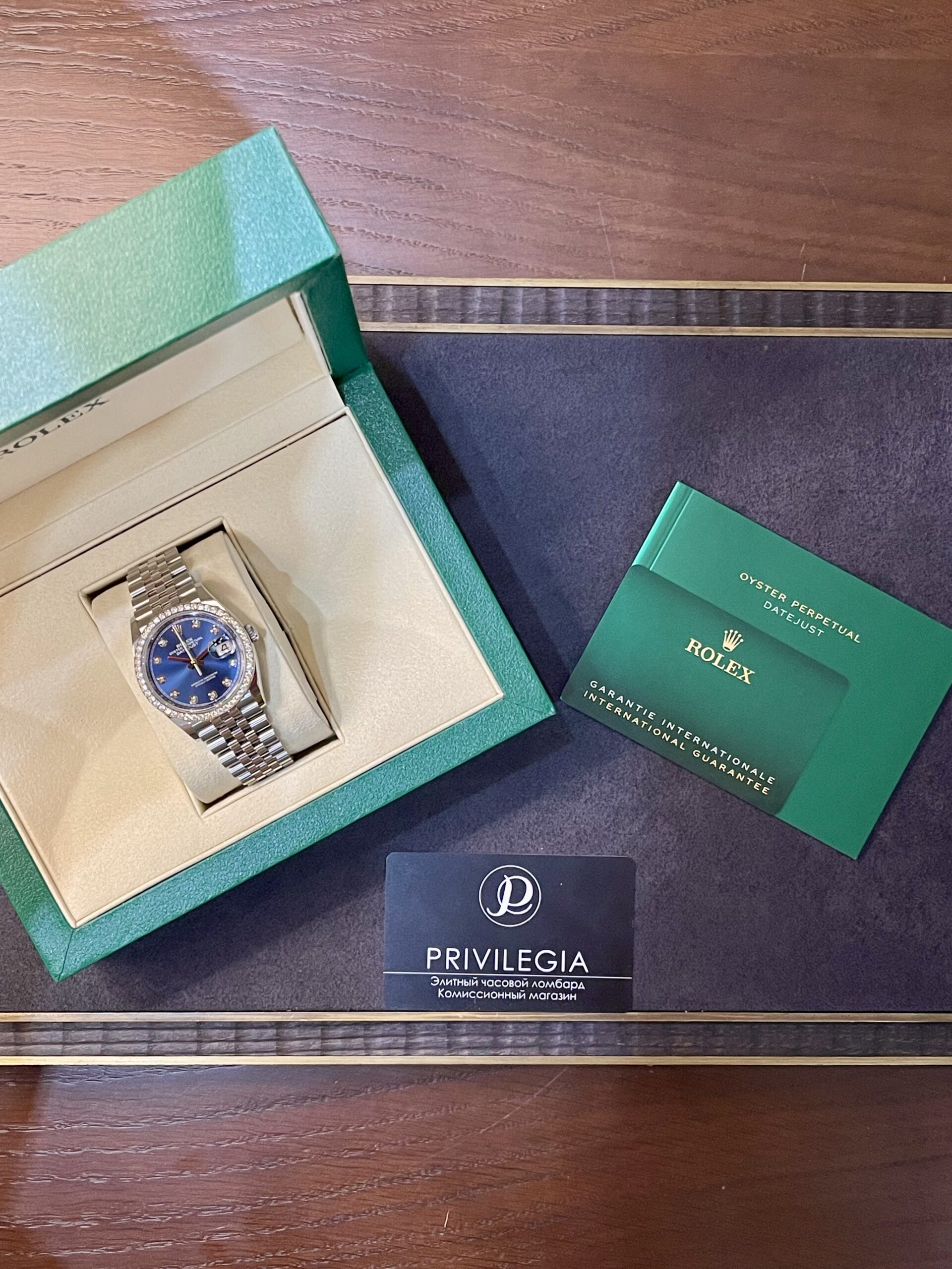 Rolex wristwatch in a turquoise box with cream interior, plus green warranty booklet and Privilegia card on a dark surface.