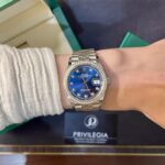 Wrist wearing a silver Rolex Datejust with a blue dial and diamond-studded bezel, seen beside a green box and Privilegia card in the background.