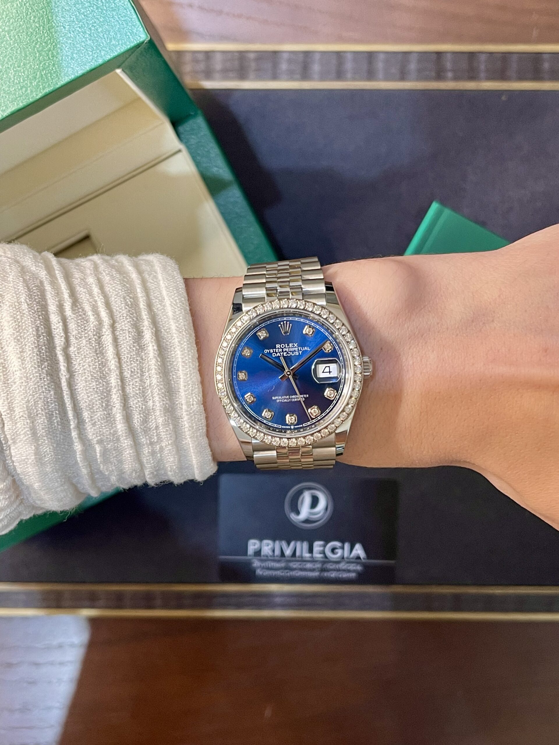 Wrist wearing a silver Rolex Datejust with a blue dial and diamond-studded bezel, seen beside a green box and Privilegia card in the background.