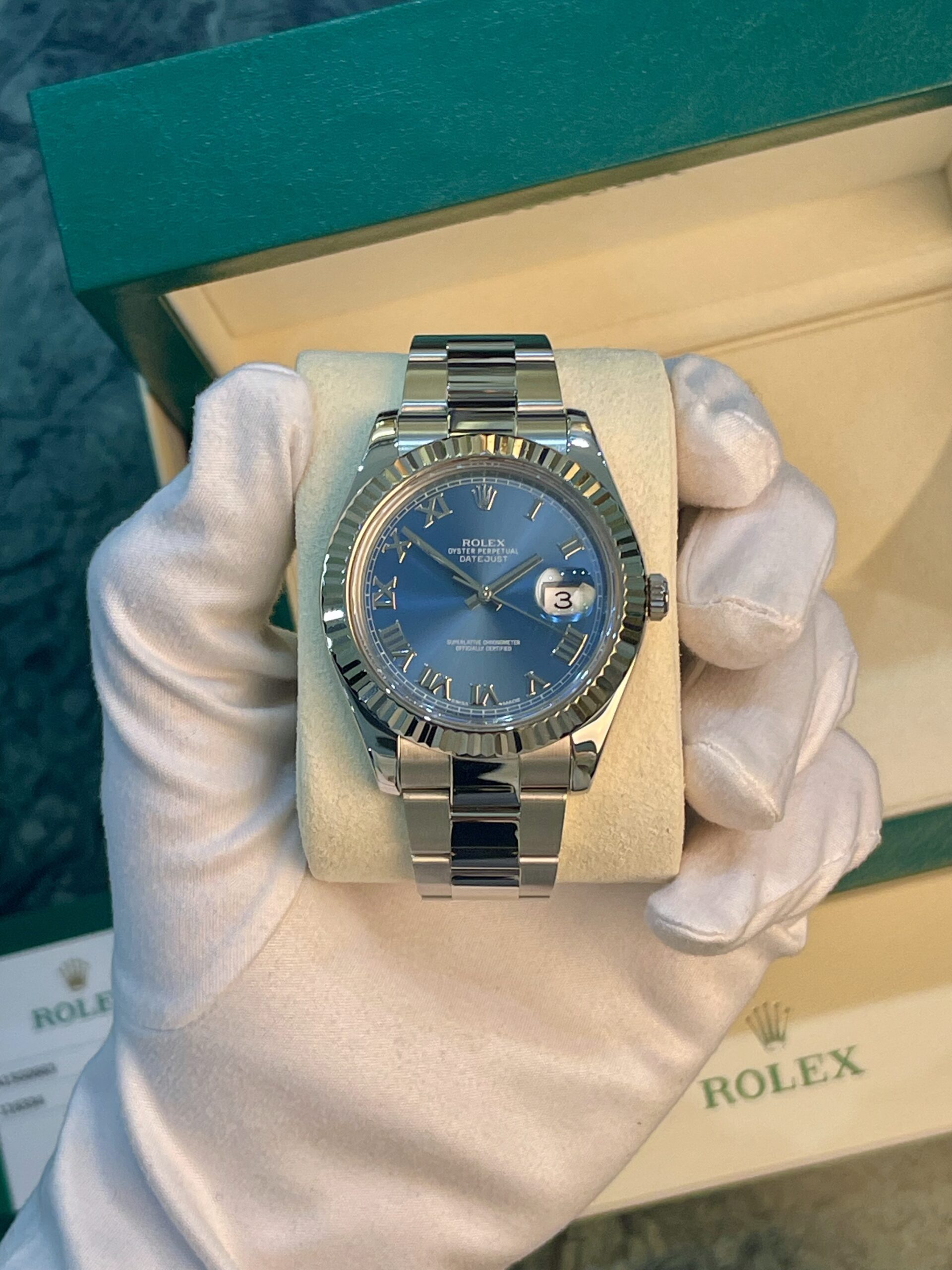 Rolex Oyster Perpetual Datejust stainless steel watch with a blue dial and date window, held by gloved hands in a display box
