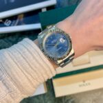Silver Rolex Datejust on a wrist with a blue dial, Roman numerals, and date at 3 o'clock.
