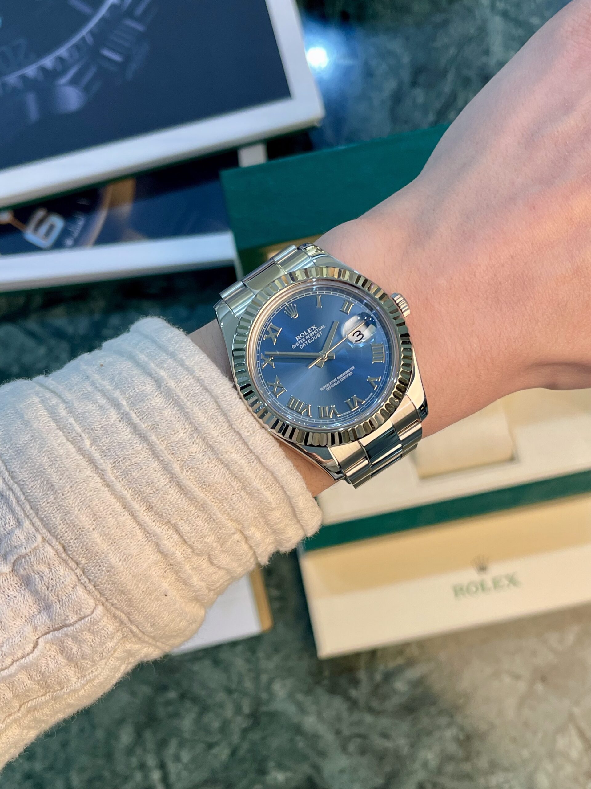 Silver Rolex Datejust on a wrist with a blue dial, Roman numerals, and date at 3 o'clock.