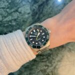 Wrist wearing a TAG Heuer Aquaracer stainless steel watch with a green bezel, beige sweater sleeve, on a rocky outdoor surface.