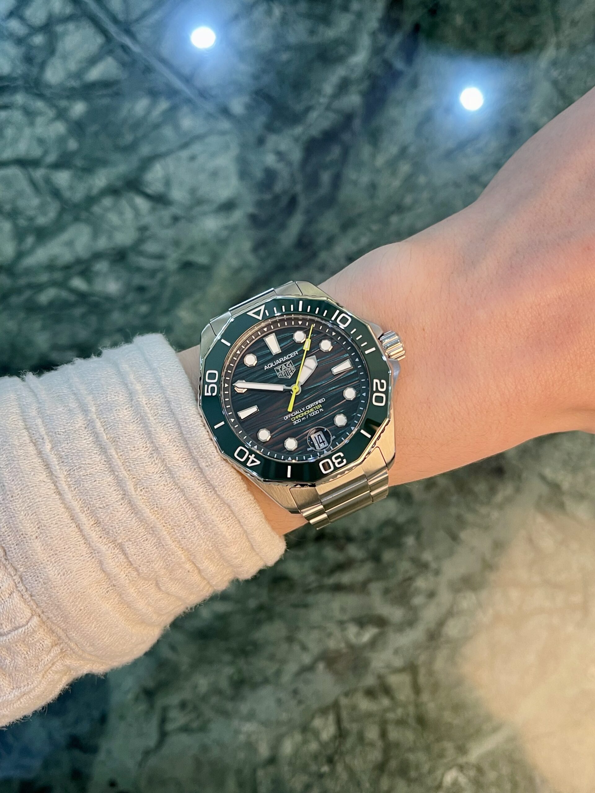 Wrist wearing a TAG Heuer Aquaracer stainless steel watch with a green bezel, beige sweater sleeve, on a rocky outdoor surface.