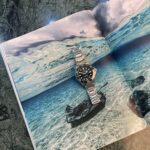 Stainless steel dive watch resting on an open magazine page showing a turquoise sea scene overlaid on rocky background