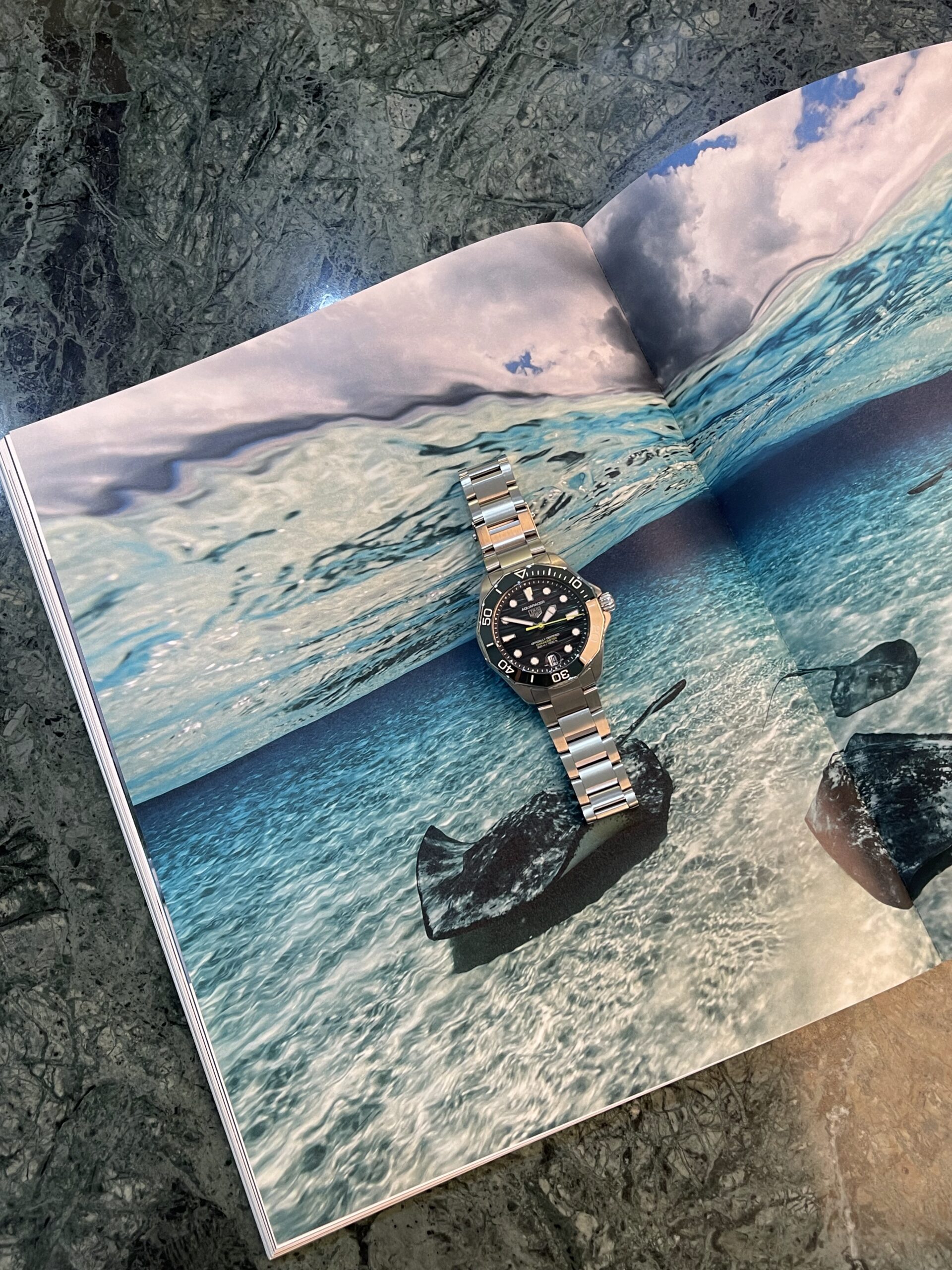 Stainless steel dive watch resting on an open magazine page showing a turquoise sea scene overlaid on rocky background