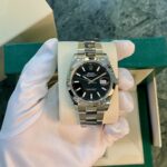 Stainless steel Rolex Datejust on a foam display, held by a gloved hand with a green Rolex box in the background.