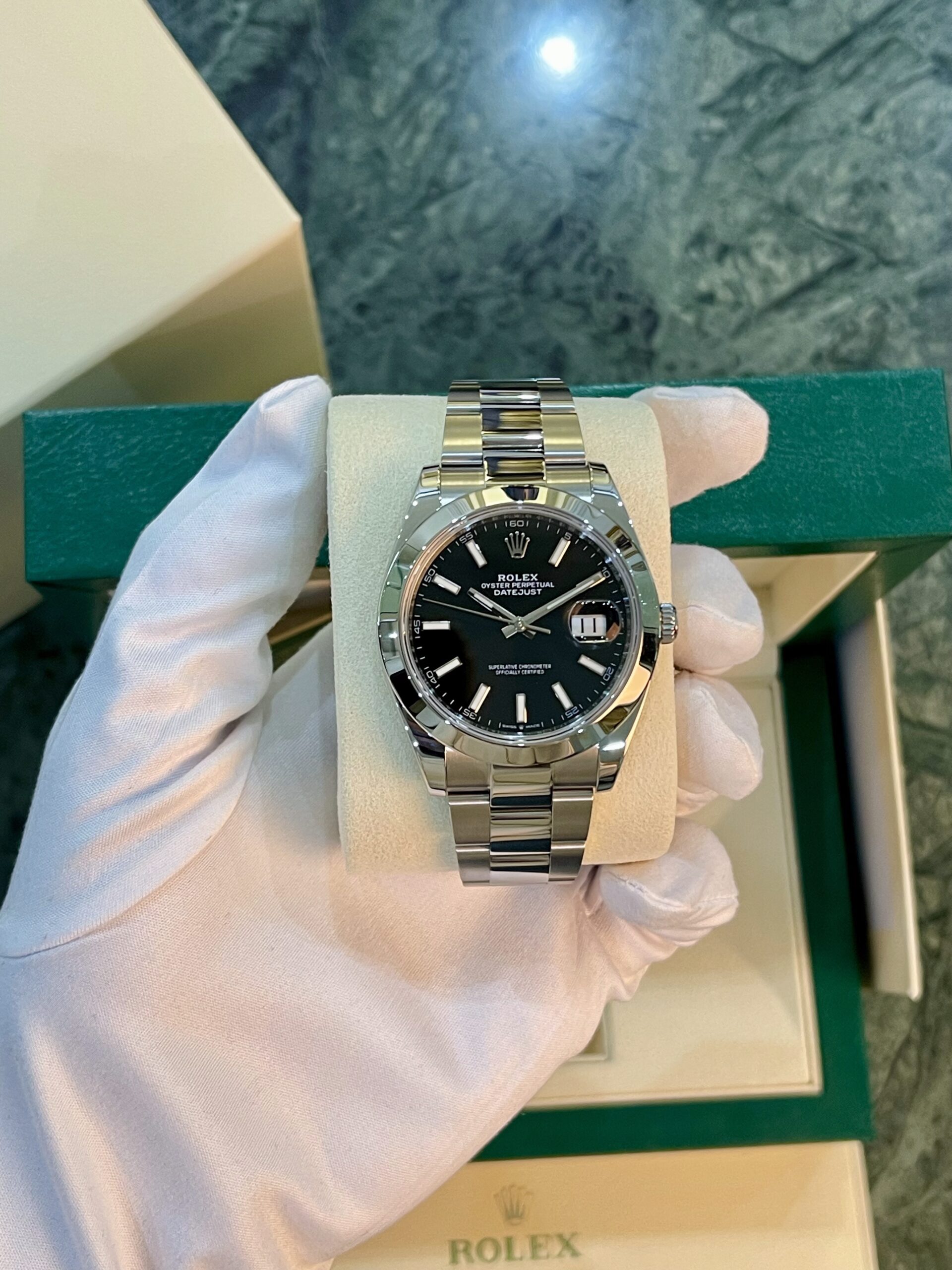 Stainless steel Rolex Datejust on a foam display, held by a gloved hand with a green Rolex box in the background.