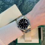 Wrist wearing a stainless steel Rolex Datejust with black dial and date window; box in background.