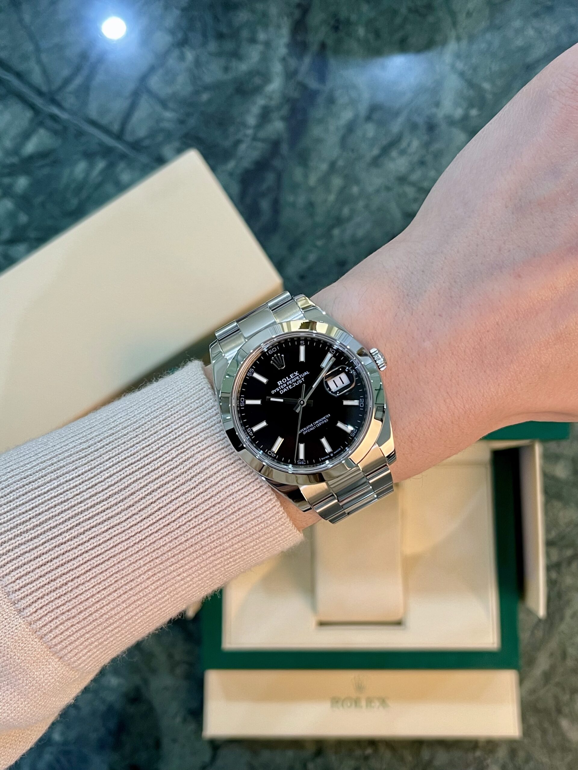 Wrist wearing a stainless steel Rolex Datejust with black dial and date window; box in background.