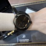 Black round wristwatch with a black strap worn on the left wrist, resting inside an open presentation box with Privilegia branding visible.