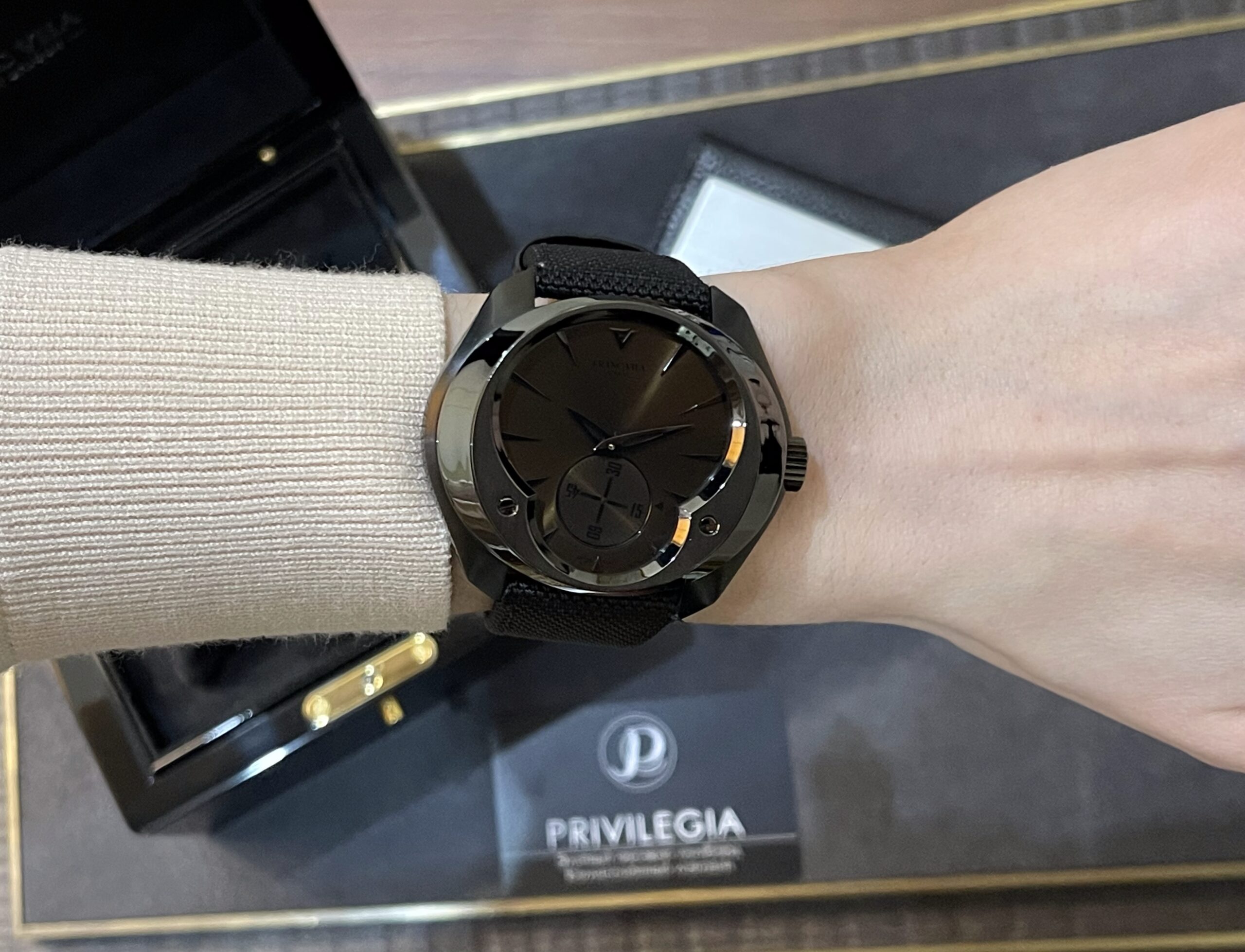 Black round wristwatch with a black strap worn on the left wrist, resting inside an open presentation box with Privilegia branding visible.