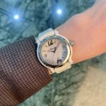 White wristwatch with a white strap on a brown textured sleeve, against a blue-green marble background.