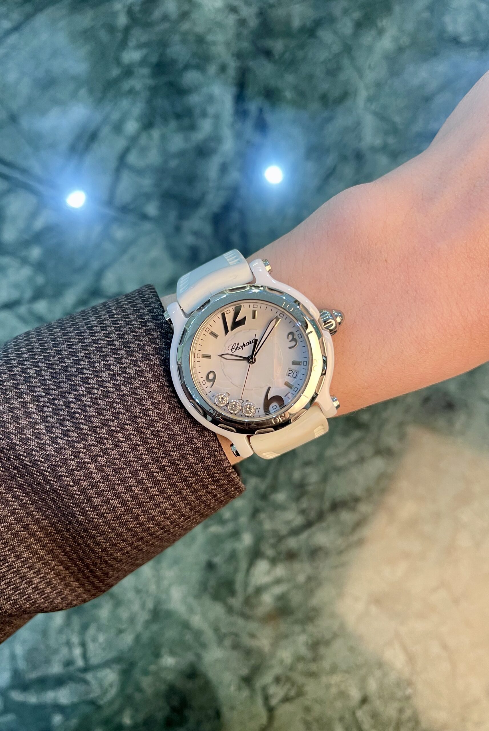 White wristwatch with a white strap on a brown textured sleeve, against a blue-green marble background.