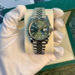 Rolex Datejust stainless steel watch with a green dial on a beige cushion, held by gloved hands in a green box setting.