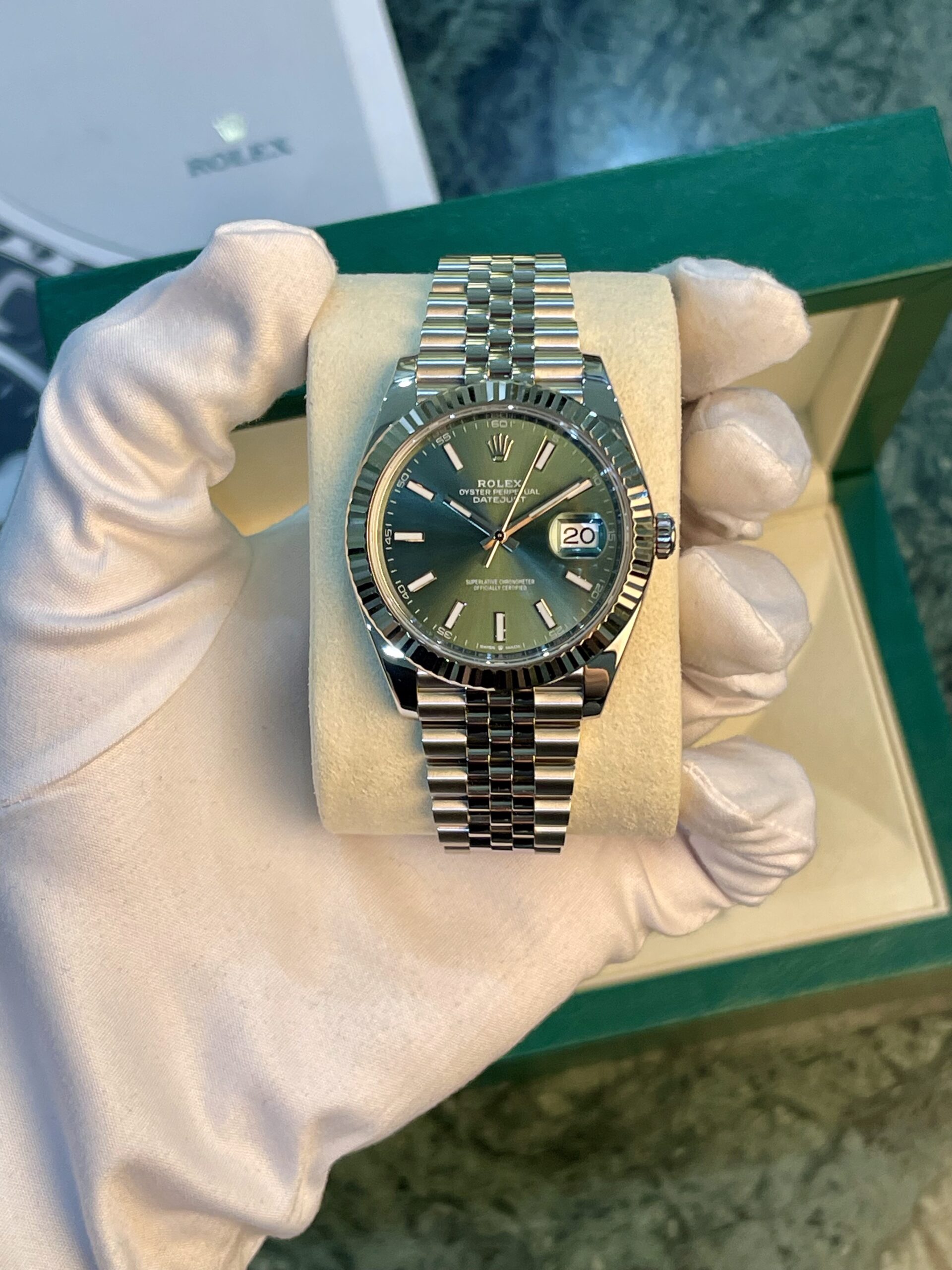 Rolex Datejust stainless steel watch with a green dial on a beige cushion, held by gloved hands in a green box setting.