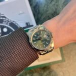 Gold and steel Rolex wristwatch with green dial on a brown textured sleeve, date visible at 3 o'clock, Jubilee bracelet, background shows a blurred box.