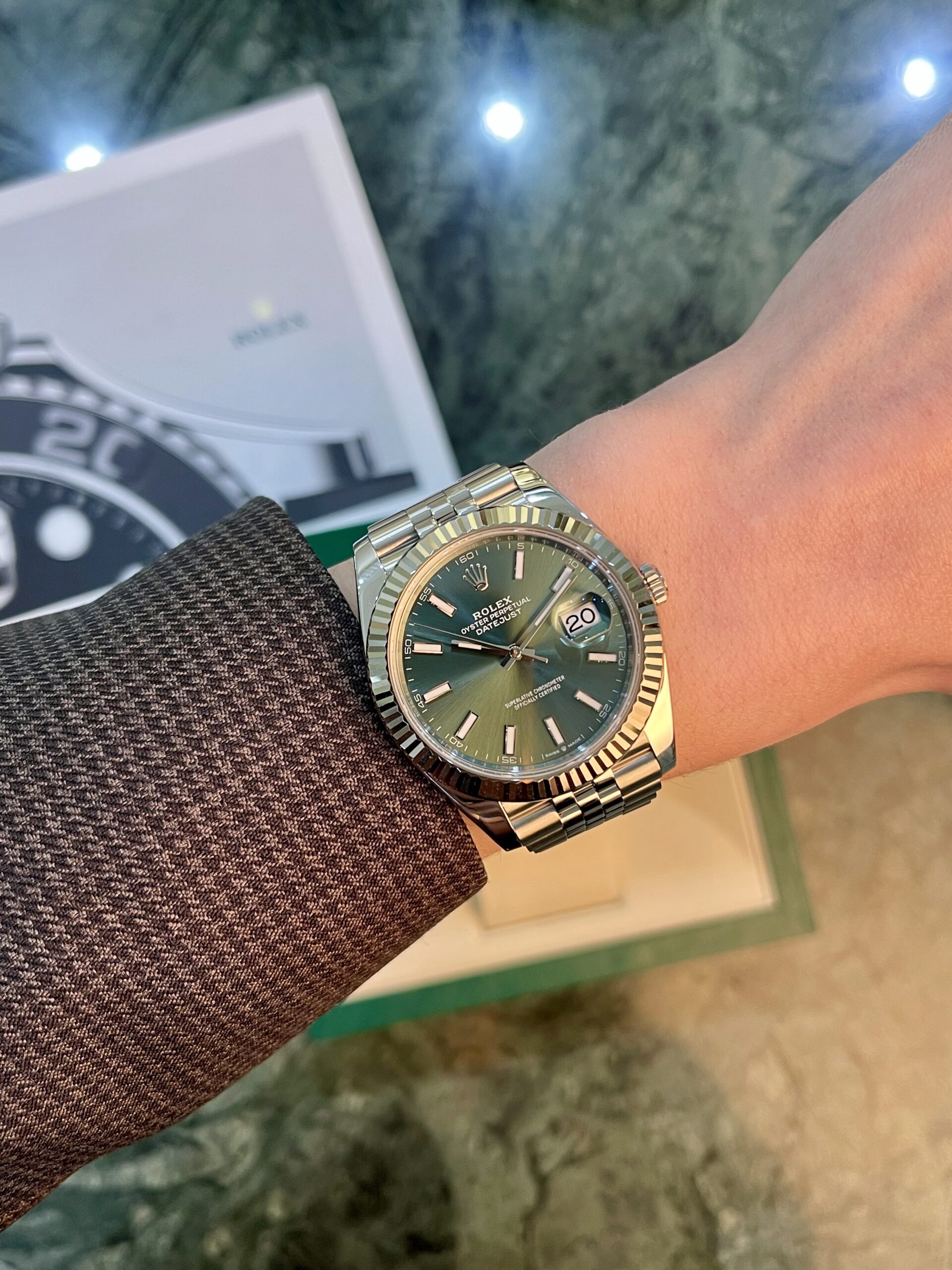 Gold and steel Rolex wristwatch with green dial on a brown textured sleeve, date visible at 3 o'clock, Jubilee bracelet, background shows a blurred box.