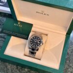 Rolex watch in its green presentation box with beige interior and warranty card nearby on a marble surface, showing a black dial and date window.
