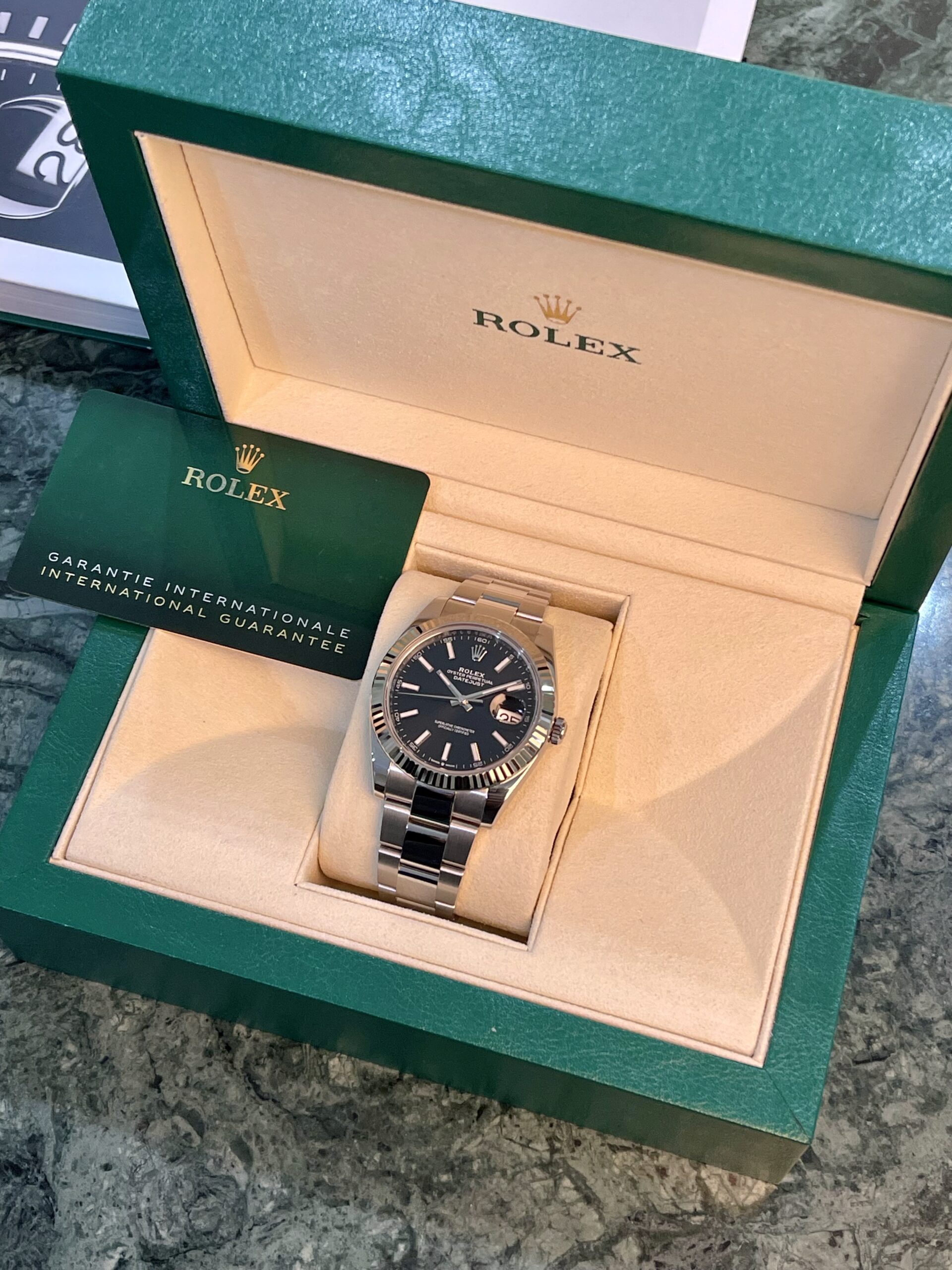 Rolex watch in its green presentation box with beige interior and warranty card nearby on a marble surface, showing a black dial and date window.